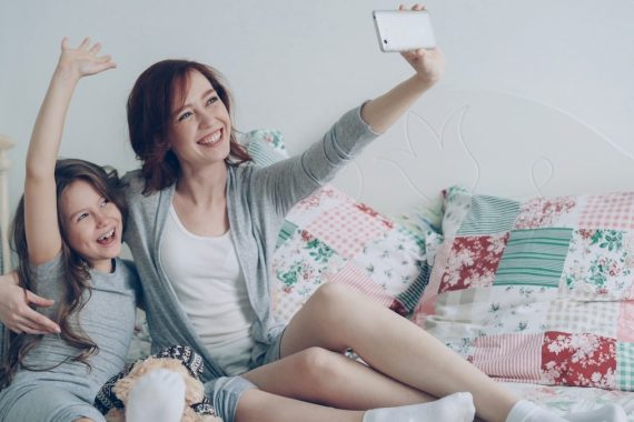 Happy mother and little girl taking selfie photo with smartphone camera and have fun grimacing while sitting in cozy bedroom at home